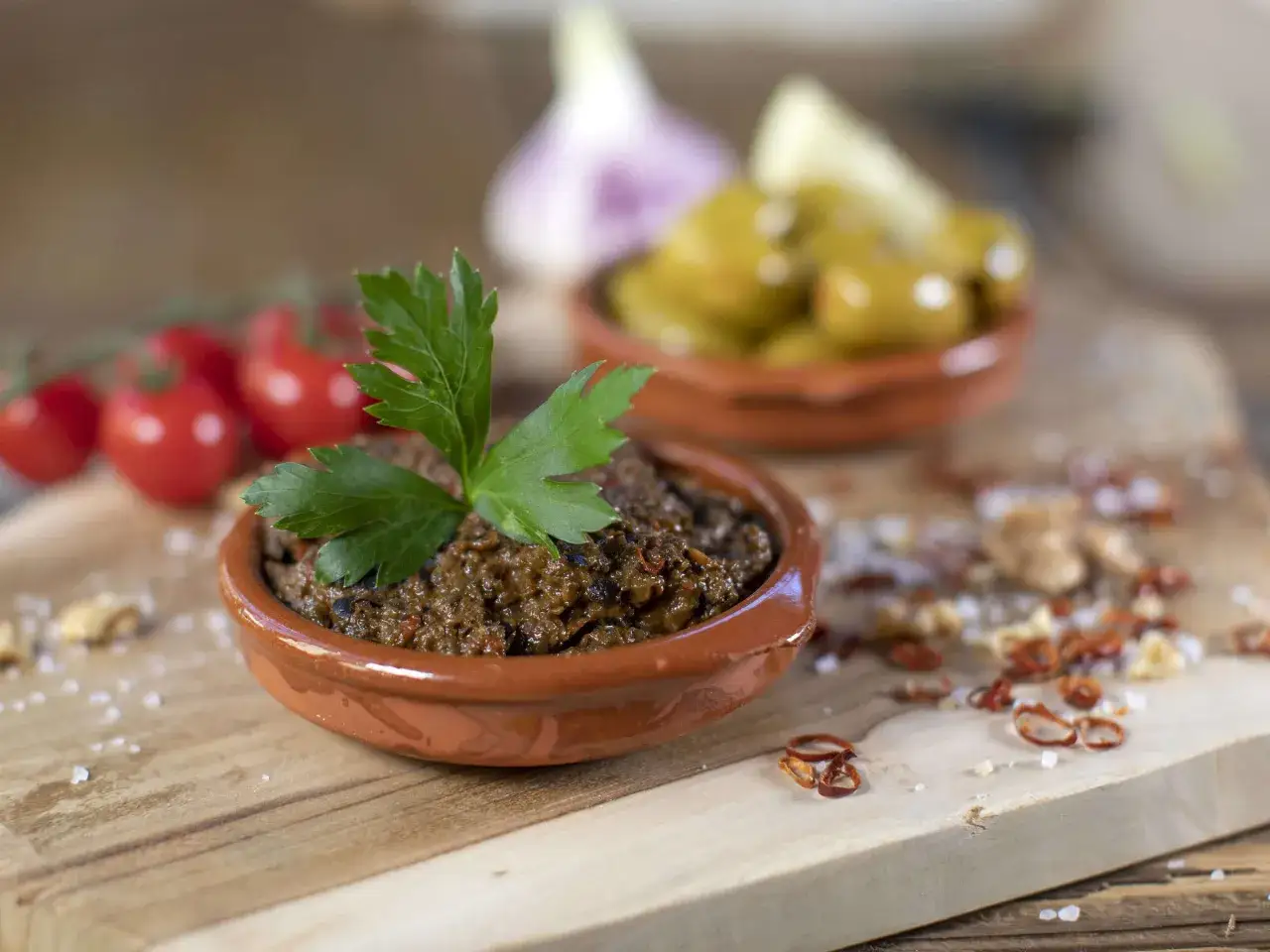 Tapenade-Rezept: Olivenpaste in Tonschale, garniert mit Petersilie. Im Hintergrund Kirschtomaten, Knoblauch und gr&uuml;ne Oliven.