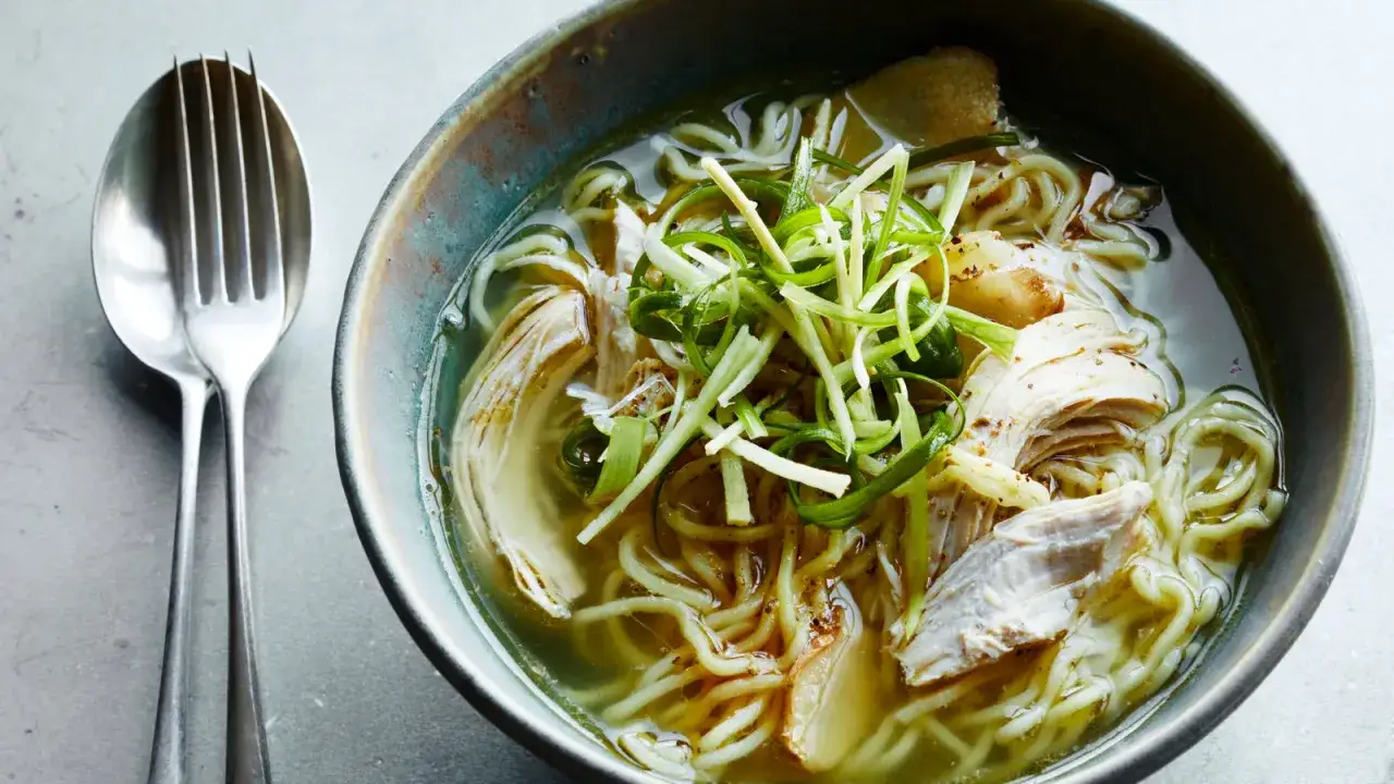 Eine dampfende asiatische Suppe mit Huhn, Nudeln und Fr&uuml;hlingszwiebeln in einer rustikalen Sch&uuml;ssel, daneben ein L&ouml;ffel und eine Gabel.