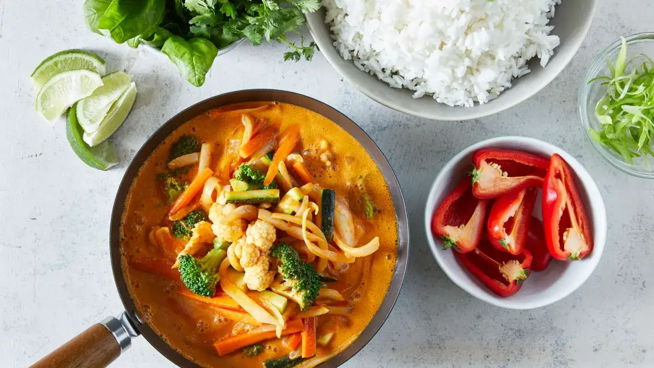 Ein dampfender roter Thai Curry mit viel Gem&uuml;se wie Brokkoli, Karotten und Zucchini, serviert mit Reis und frischen Limetten.