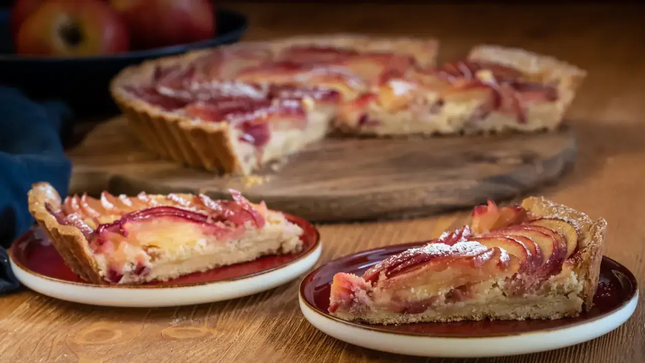 Zwei St&uuml;cke einer k&ouml;stlichen Tarte au Pomme, best&auml;ubt mit Puderzucker, auf roten Tellern. Im Hintergrund eine ganze Tarte.