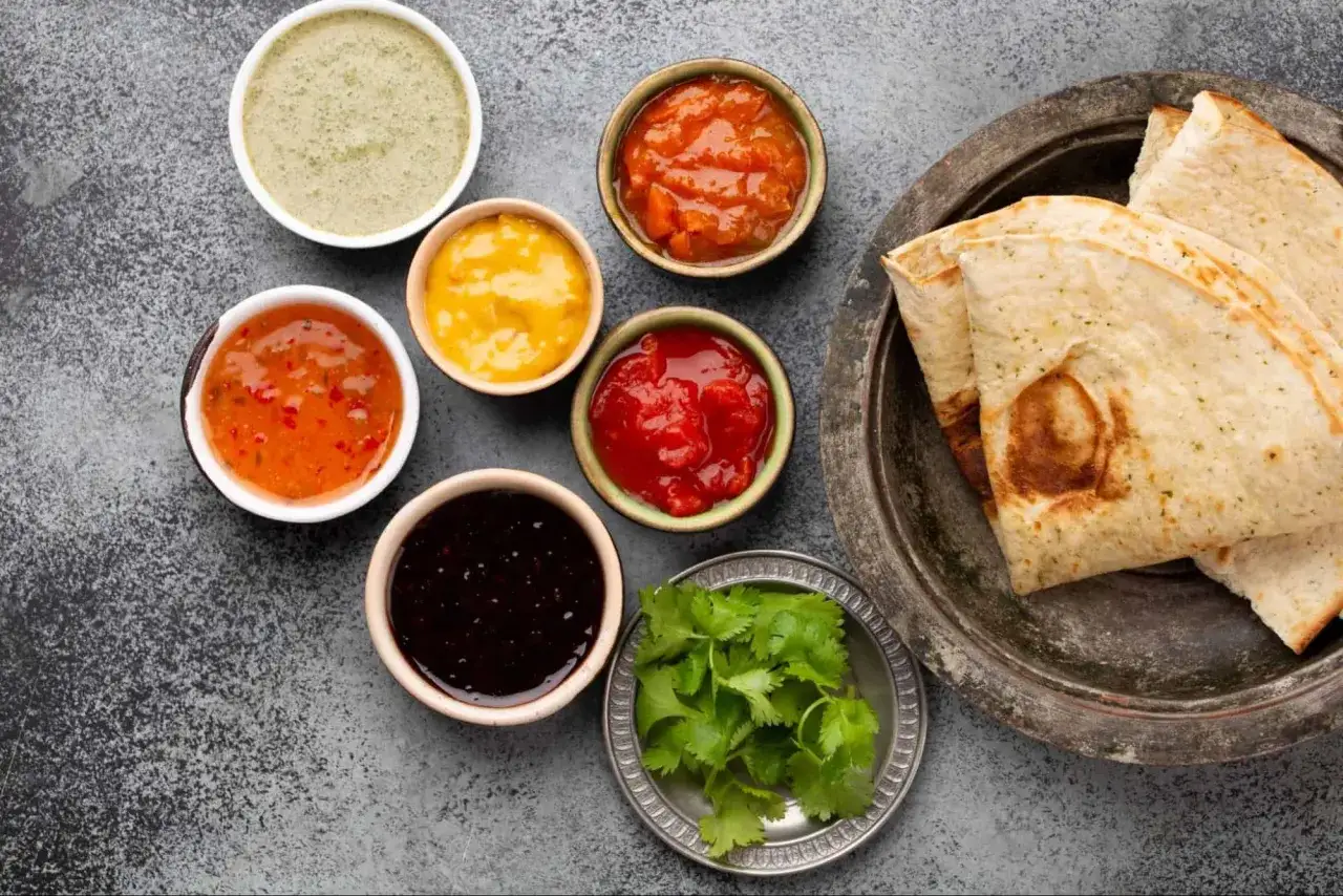 Sechs Sch&auml;lchen mit verschiedenen Saucen, darunter ein gr&uuml;nes, ein gelbes, ein orangefarbenes und ein rotes, sowie ein dunkles und ein helles Chutney. Dazu Fladenbrot und Koriander.