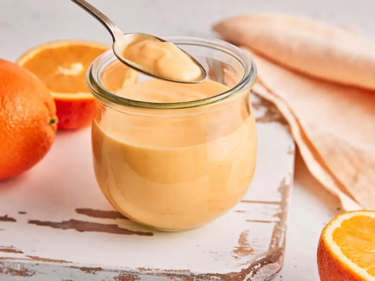 Ein L&ouml;ffel hebt cremiges orange curd aus einem Glas. Frische Orangen liegen daneben.