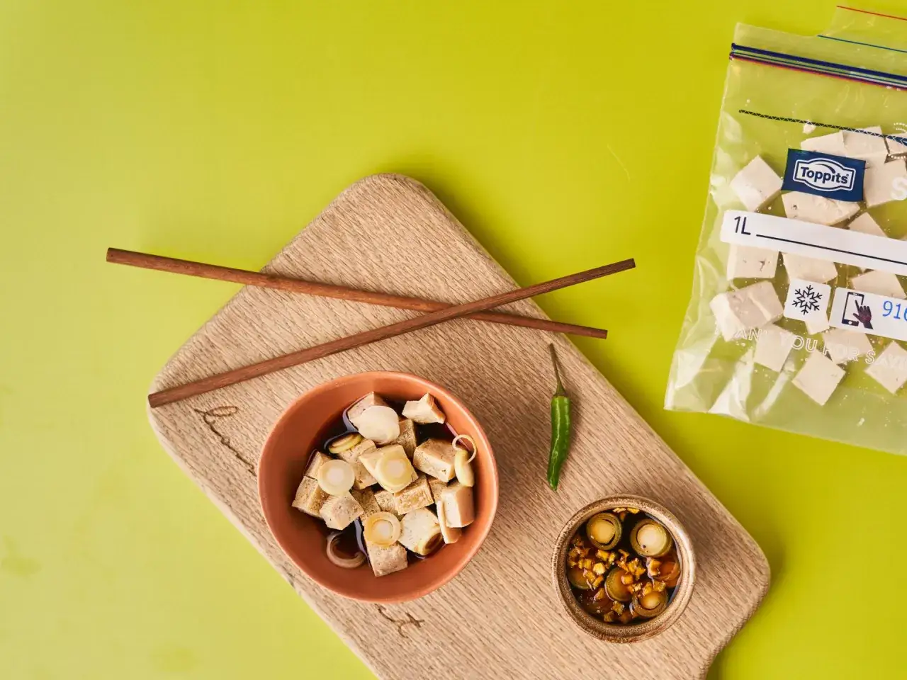 W&uuml;rfel von Tofu in einer Sch&uuml;ssel mit So&szlig;e, daneben ein Beutel mit Tofu zum Einfrieren und ein gr&uuml;ner Chili.