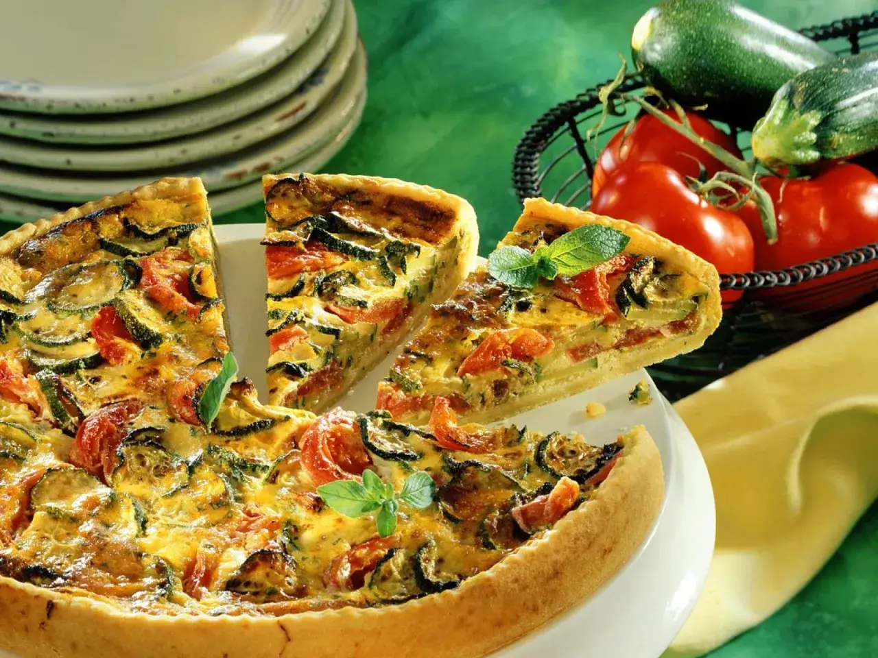 Eine k&ouml;stliche, sommerliche Quiche mit Zucchini und Tomaten, garniert mit frischen Kr&auml;utern. Daneben liegen frische Zutaten in einem Korb.