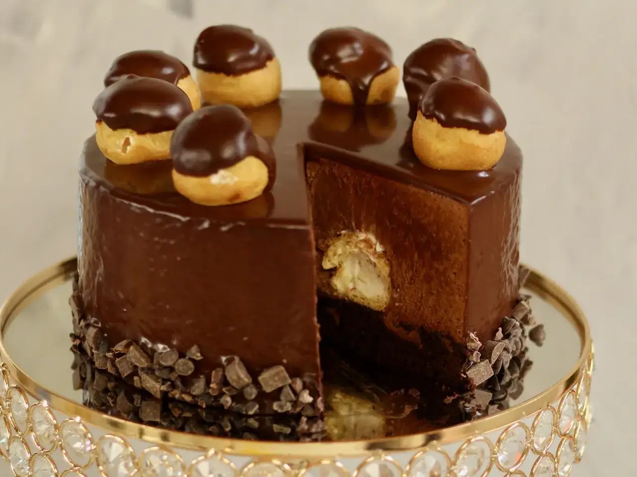 Eine glänzende Schokoladentorte, dekoriert mit kleinen Schokoladen-Profiteroles. Ein Stück ist herausgeschnitten und zeigt die cremige Füllung.