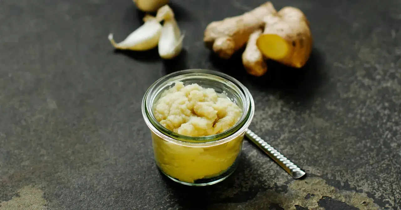 Frisch zubereitete Ingwer Knoblauch Paste in einem Glas, daneben frische Knoblauchzehen und Ingwerwurzeln auf dunklem Untergrund.