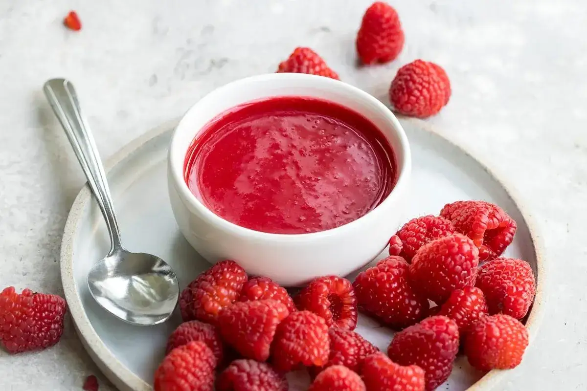 Ein Schälchen mit leuchtend rotem Himbeer-Coulis, umgeben von frischen Himbeeren und einem Löffel auf einem Teller.