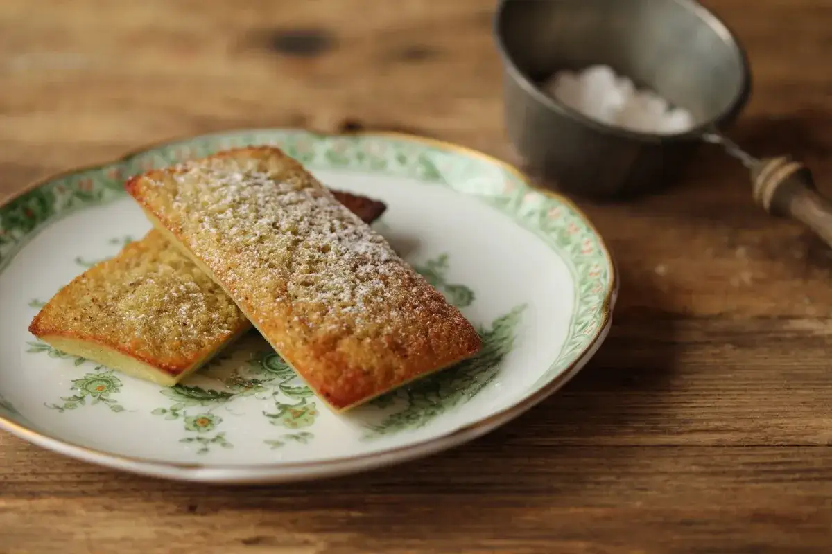 Zwei goldbraune Financiers, best&auml;ubt mit Puderzucker, auf einem Teller mit Blumenmuster. Ein L&ouml;ffel mit Zucker im Hintergrund. Ein k&ouml;stliches Financier Rezept.