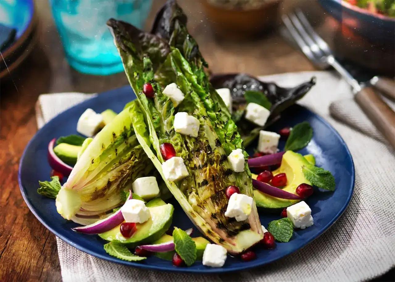 Gegrillter Romana Salat mit Avocado, Feta, Granatapfelkernen und Minze auf einem blauen Teller.
