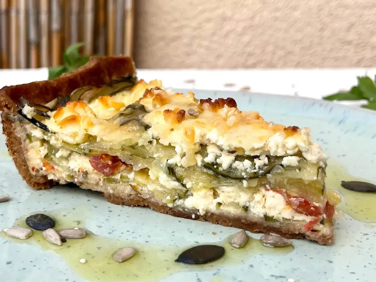 Ein St&uuml;ck herzhafte Quiche mit Zucchini, Tomaten und Feta auf einem Teller mit Kernen und &Ouml;l.