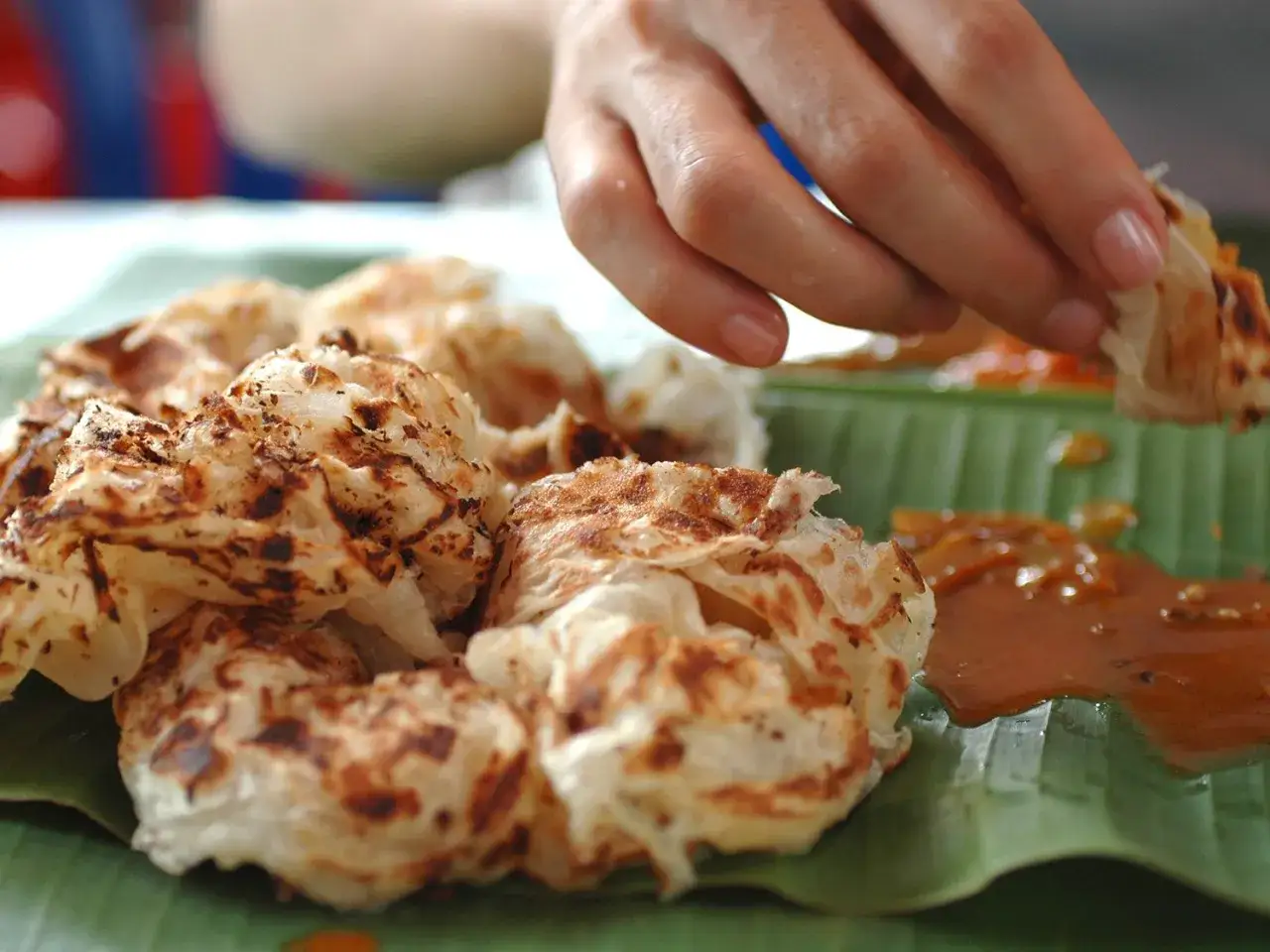 Eine Hand rei&szlig;t ein St&uuml;ck von knusprigem Roti Canai ab, das auf einem Bananenblatt mit Curry serviert wird.