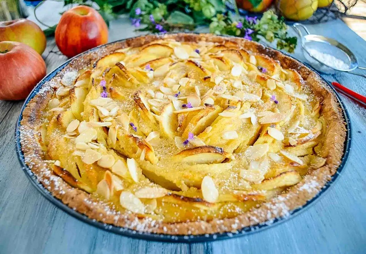 Ein goldbrauner Apfelkuchen mit Mandelbl&auml;ttchen und Puderzucker, perfekt f&uuml;r ein Apfeltarte Rezept. Daneben liegen frische &Auml;pfel.
